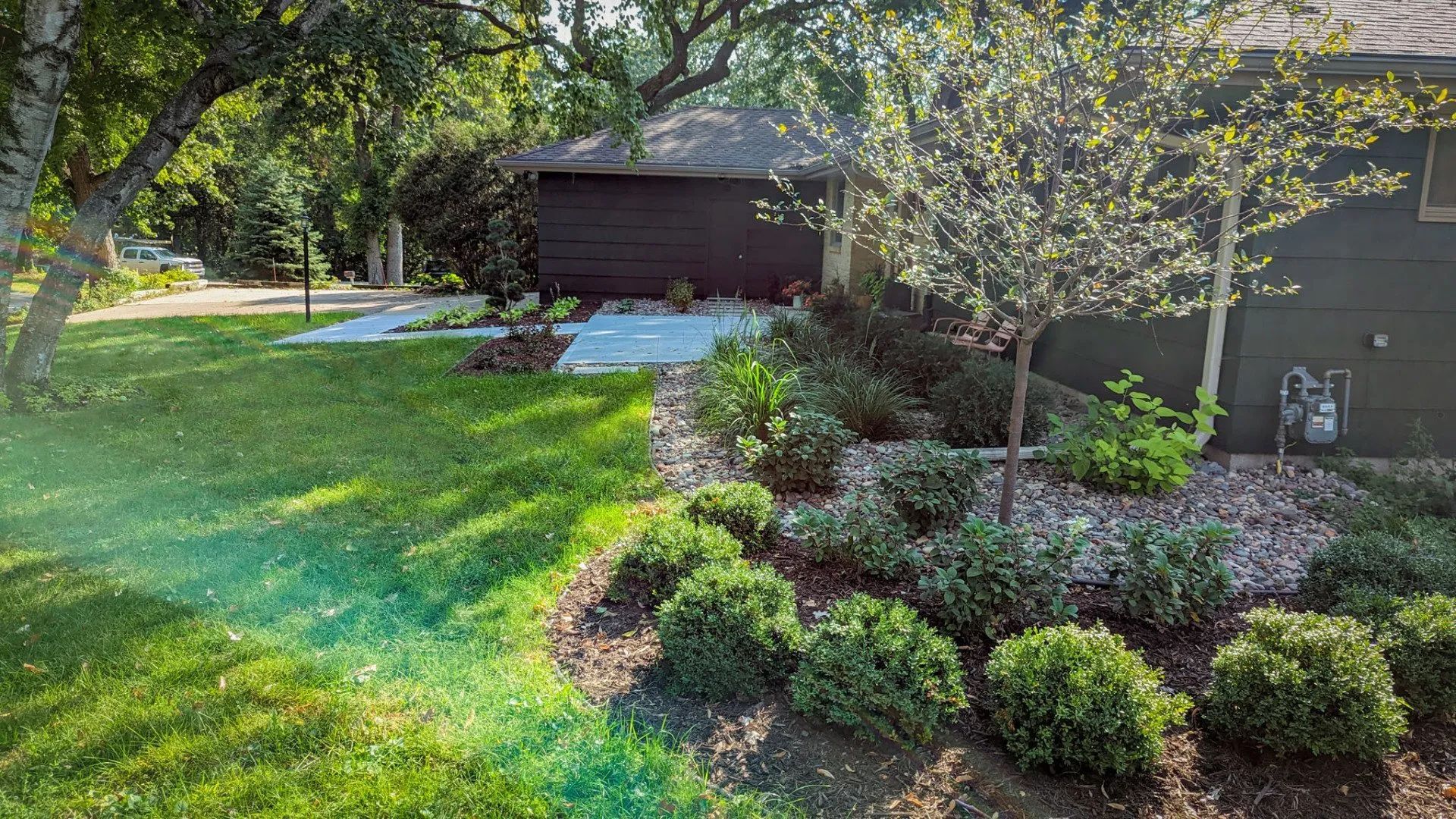 Green lawn and landscaped garden with bushes, small trees, and a dark brown garage in the background.