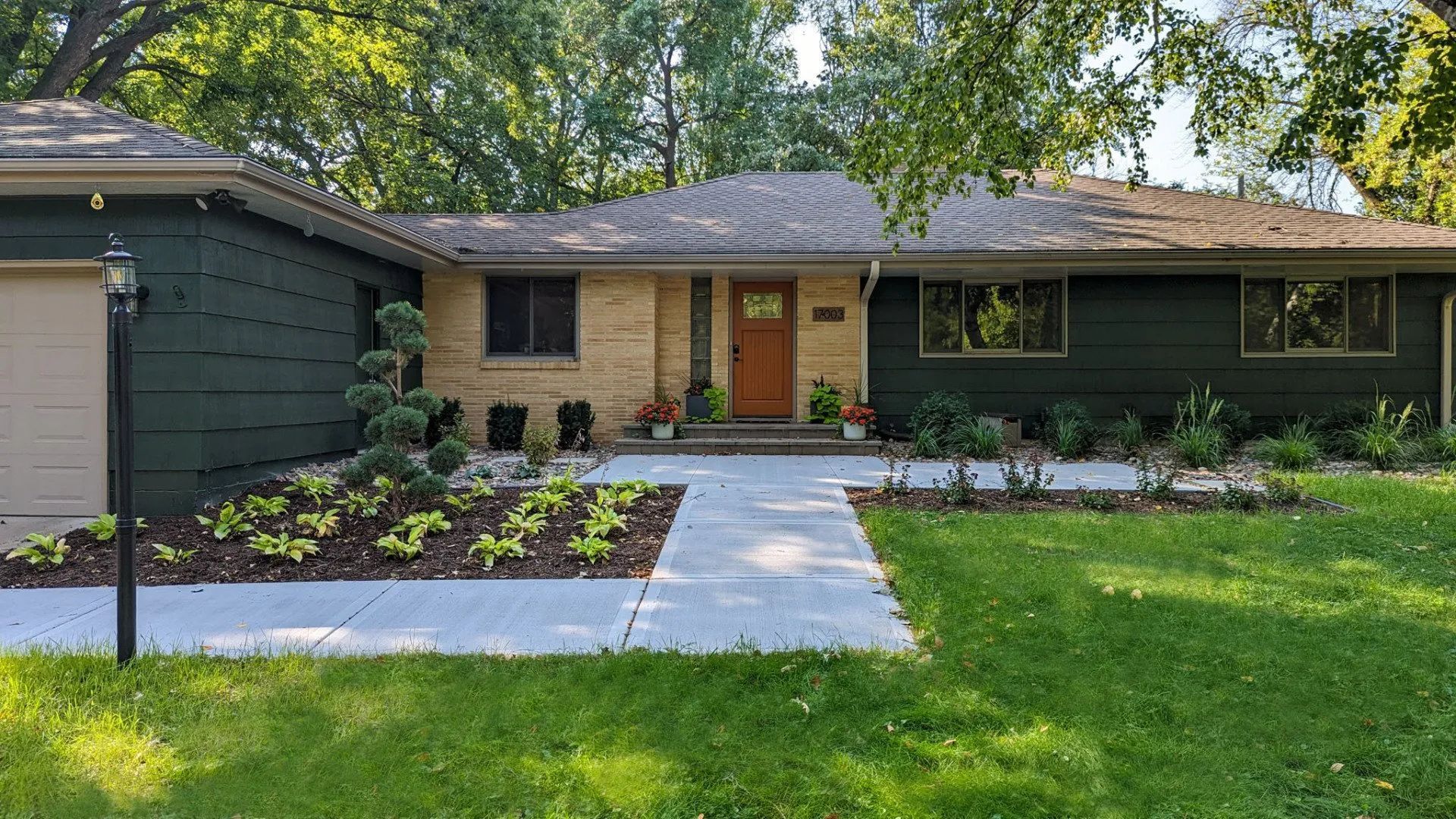 A one-story house with green siding and a brick facade. A concrete walkway leads to the front door. Landscaping and a lawn are in front.