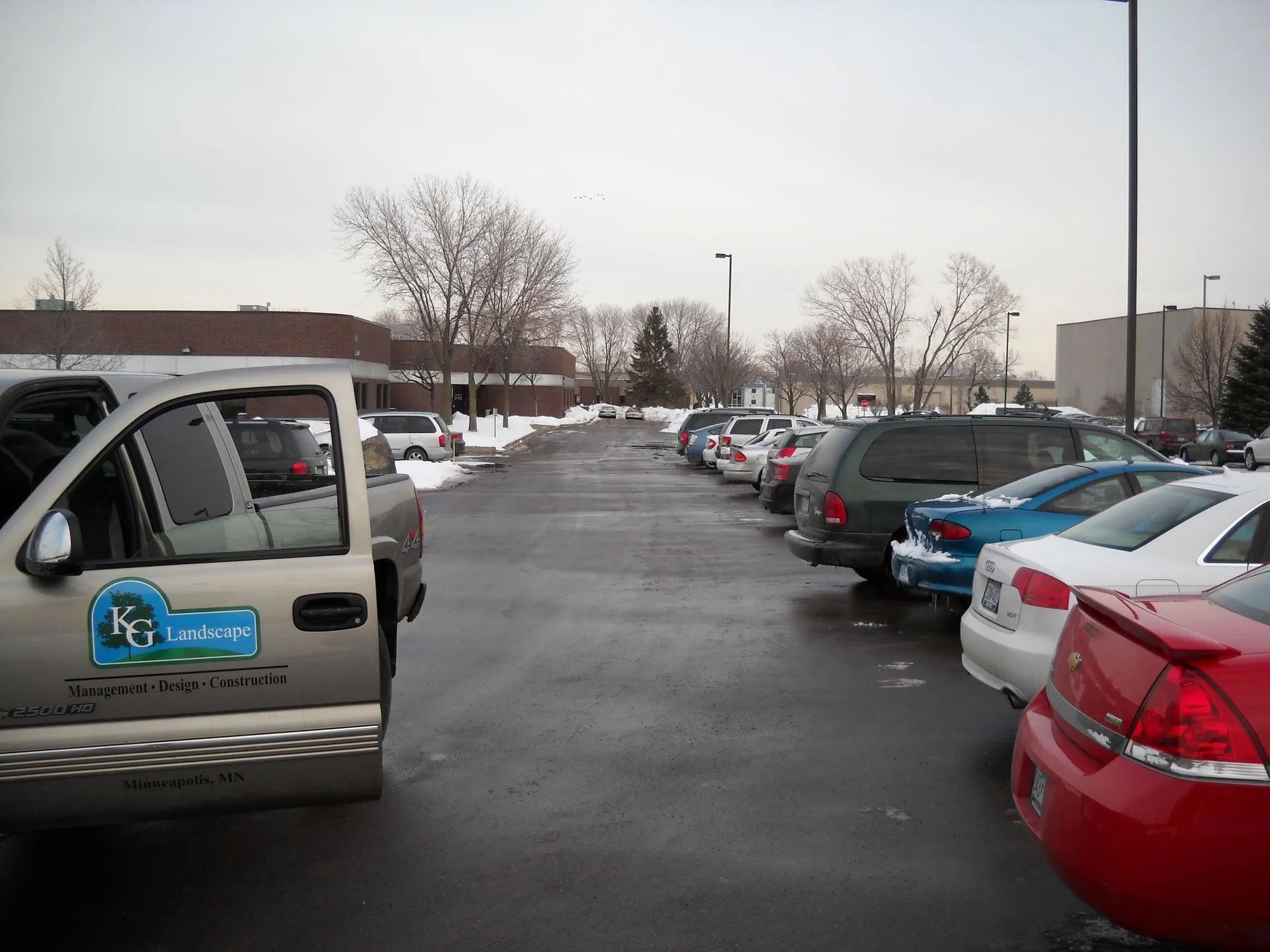 A parking lot with cars and a landscaping truck on a cloudy day, with a building in the background.
