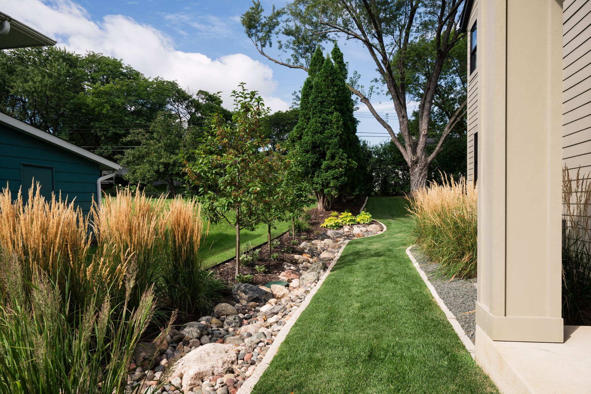 Lush green lawn with a rock garden border leading to a stone patio. A young tree is in the foreground.
