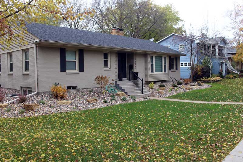 A single-story gray brick house with a dark shingled roof, stone landscaping, and a small front stoop, set in a yard.