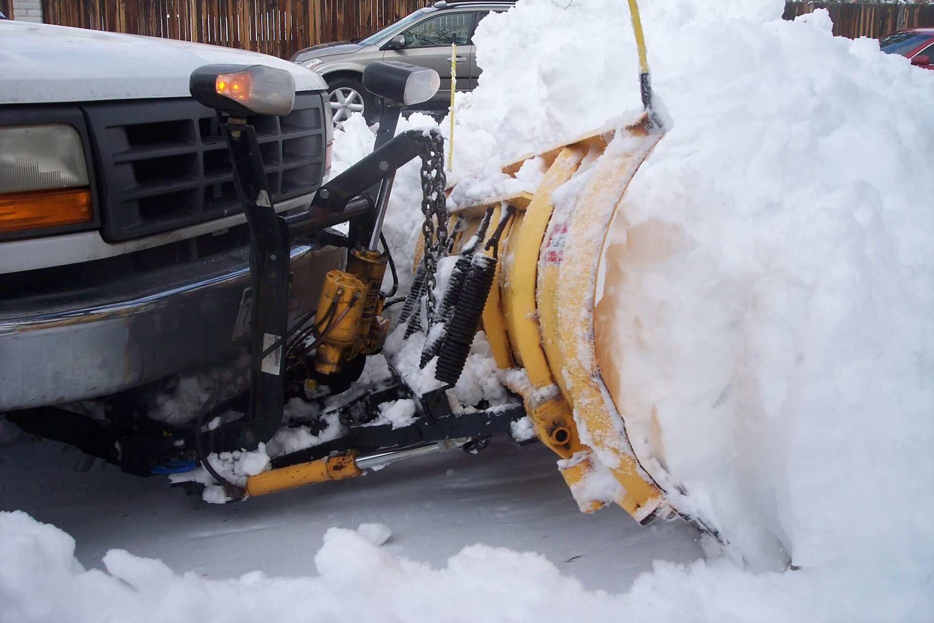A close-up view of a yellow snow plow mounted to the front of a white truck, pushing a large pile of fresh snow.