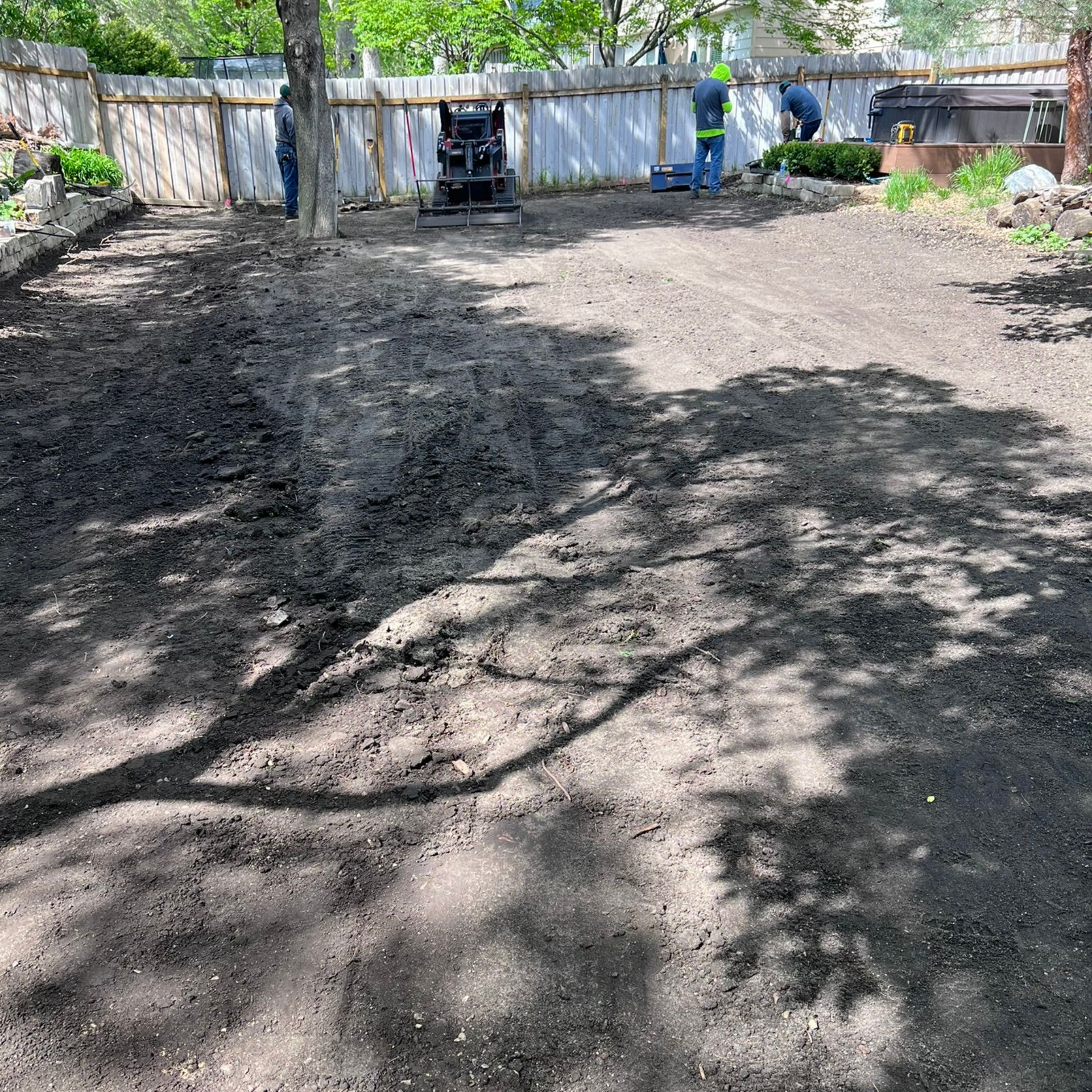 Workers in a sunny backyard clearing dirt and preparing the ground for landscaping near a wooden fence.