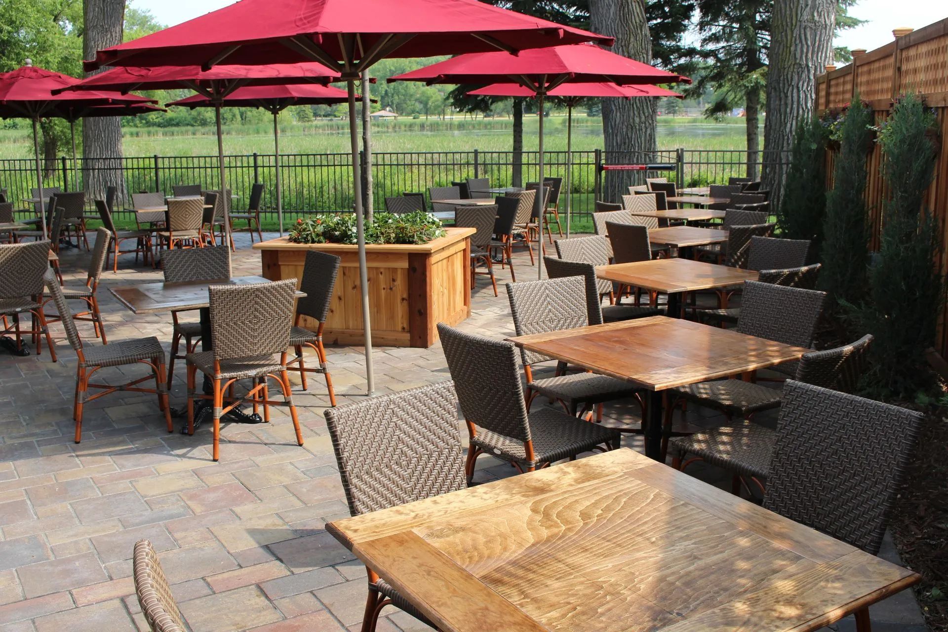 Outdoor dining patio with red umbrellas, tables, and chairs, beside a fence and trees.