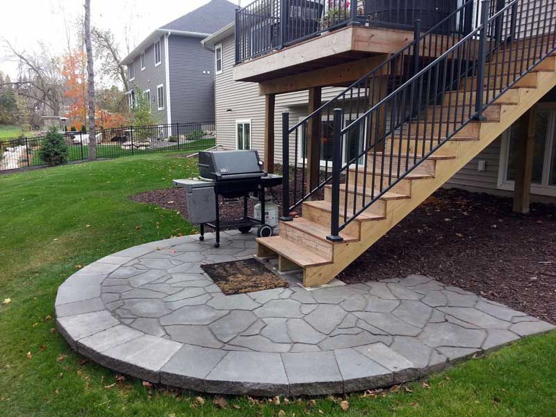 A stone patio with a curved border sits beneath a wooden deck and staircase at the back of a house with a grill.
