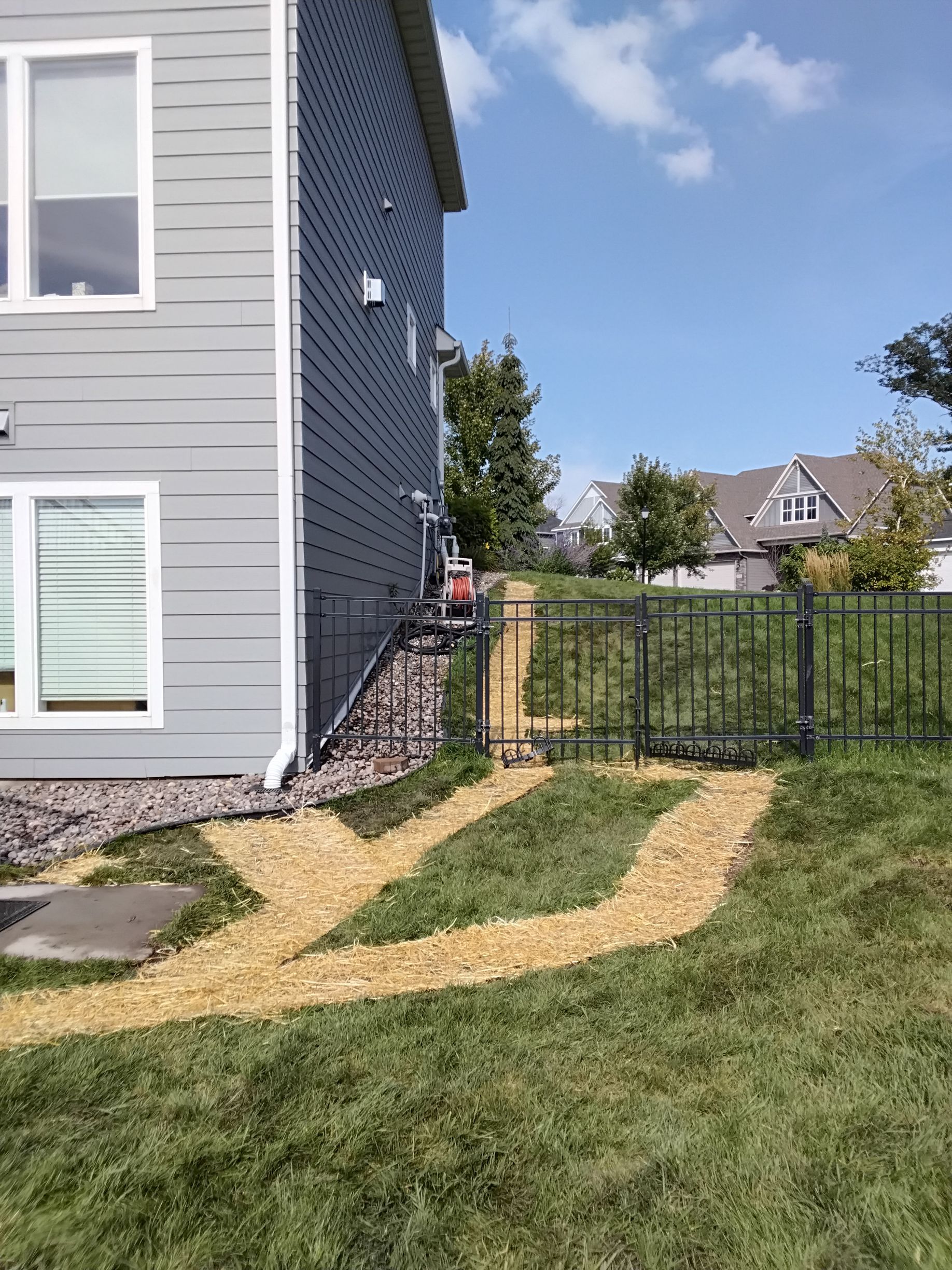 A gray house exterior with a wood-chip path leading to a metal fence gate in a grassy backyard.