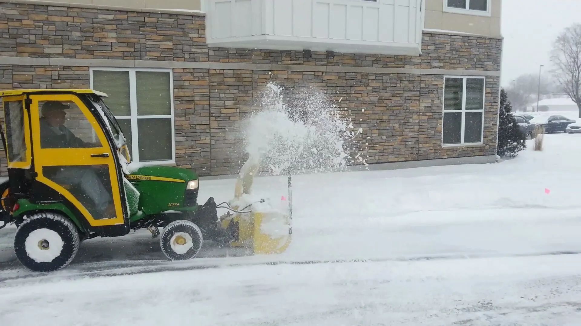 John Deere tractor with snow blower clearing snow from a paved area in front of a stone building.
