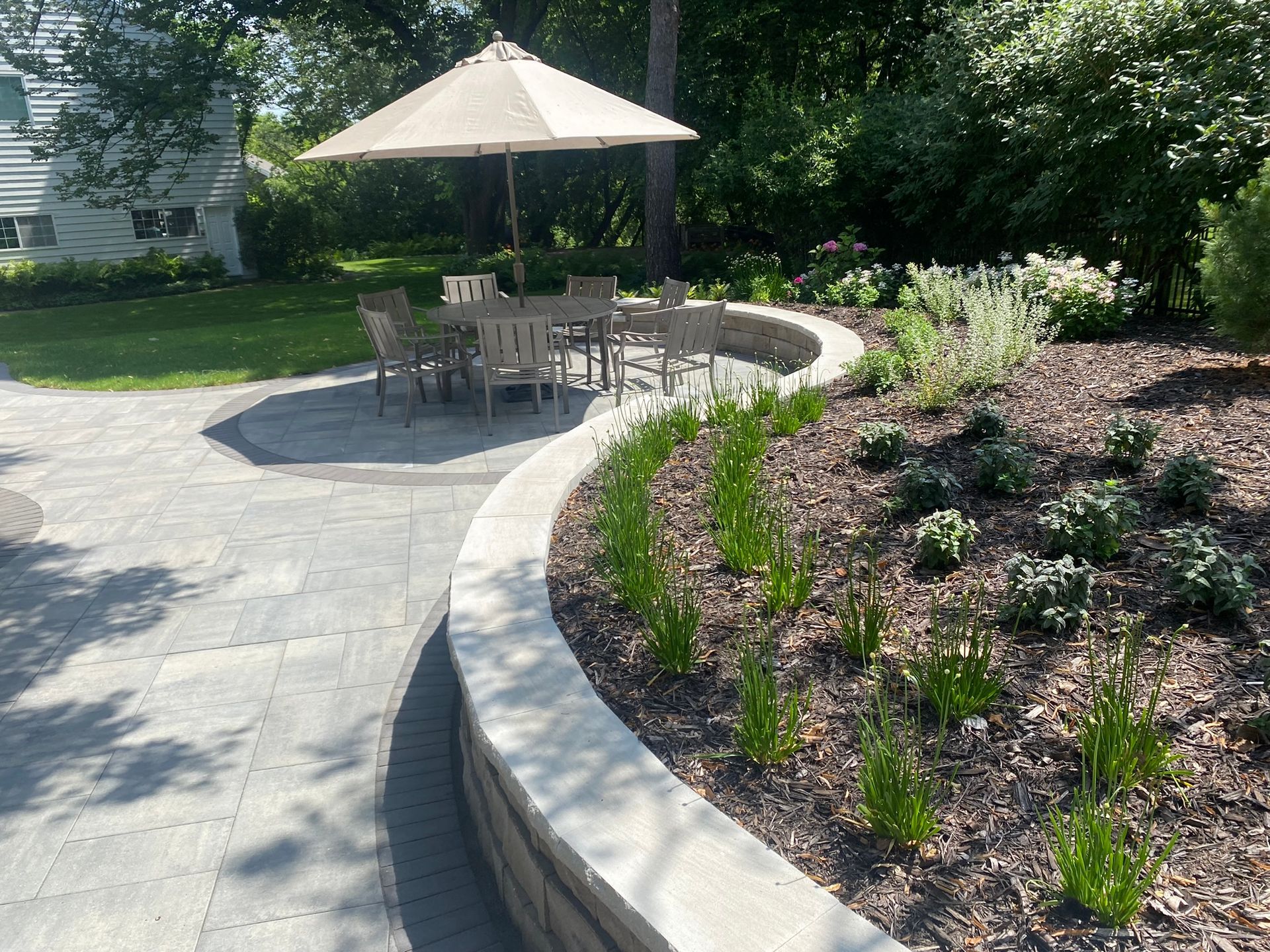 A backyard patio with a dining set under an umbrella, a curved retaining wall with plants, and a lawn.