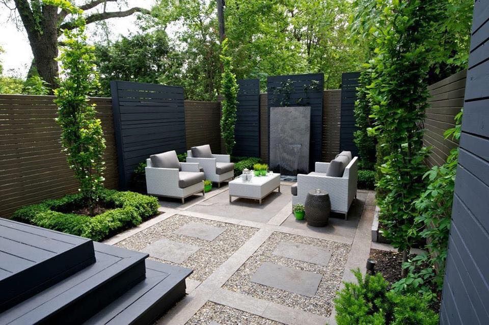 A modern outdoor patio with seating, water feature, and stone pathway surrounded by lush greenery and dark gray wooden walls.