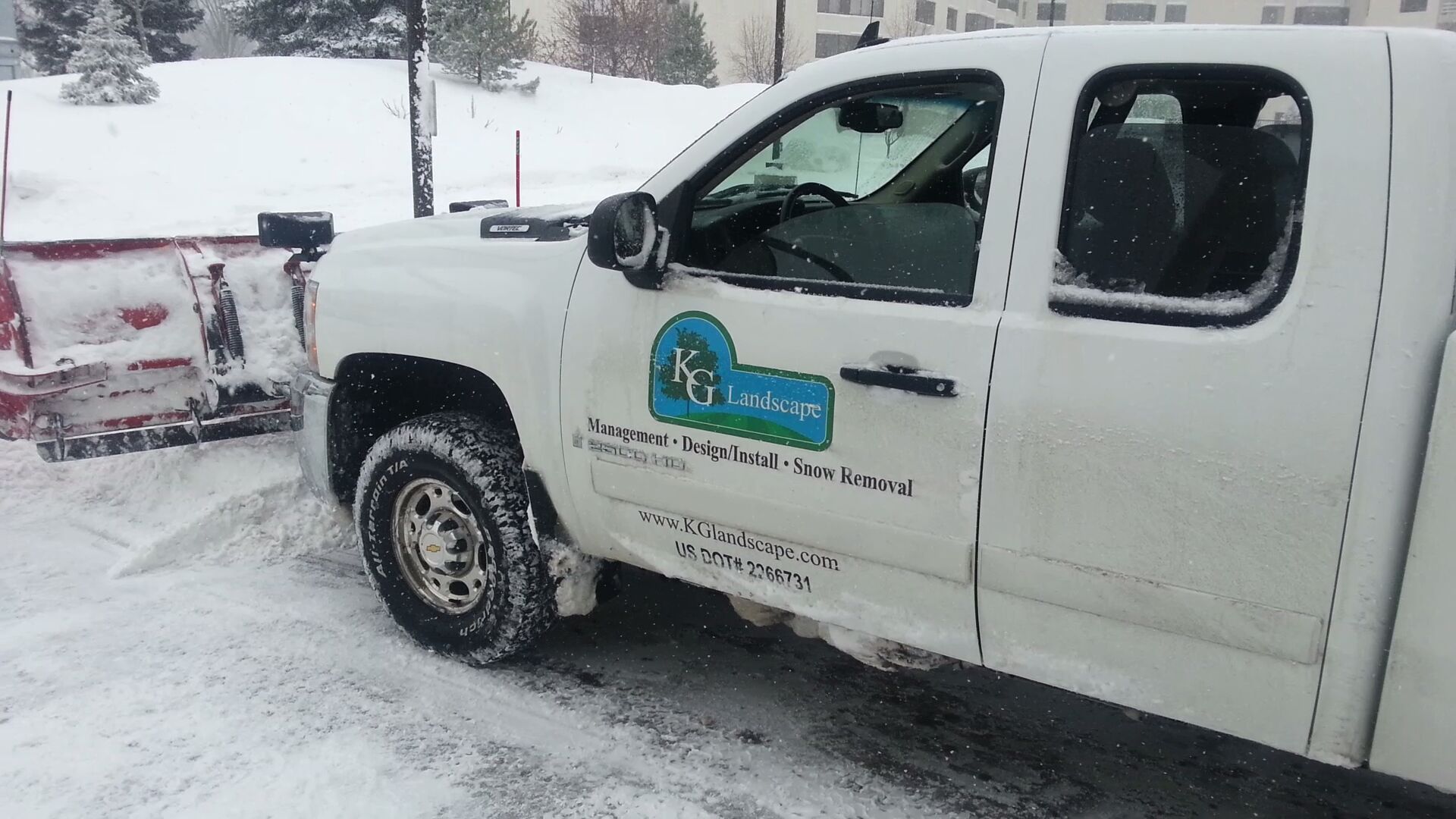 A white snow plow truck with a company logo on the door parked in a snowy parking lot.