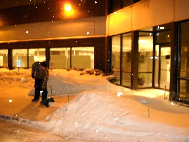 Person standing near snow-covered building entrance at night with falling snow. Yellow lights reflect.