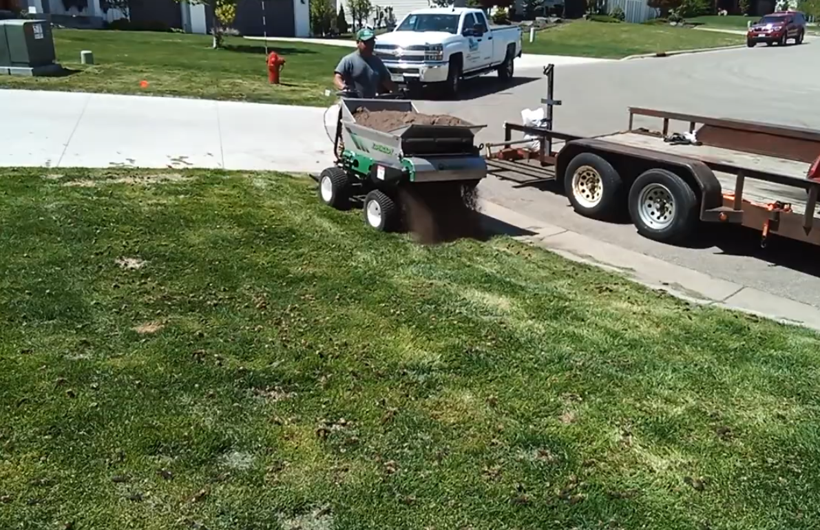 A person operates a machine to spread soil onto a green lawn near a parked truck and trailer on a sunny day.