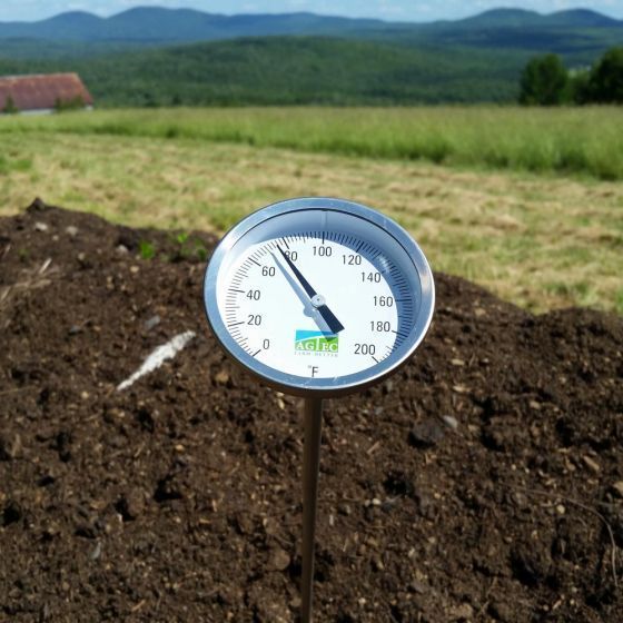Thermometer in a compost pile, outdoors. The dial reads 120 degrees Fahrenheit, with green fields and mountains in the background.