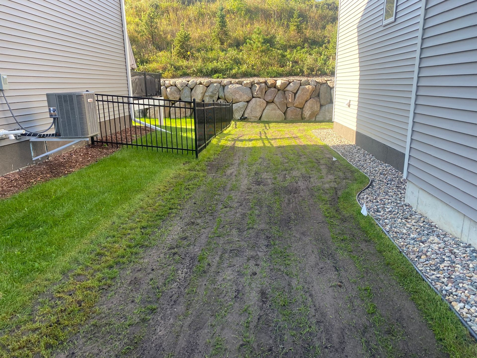 A side yard between two houses features a path of patchy, thin grass and soil leading toward a stone retaining wall.