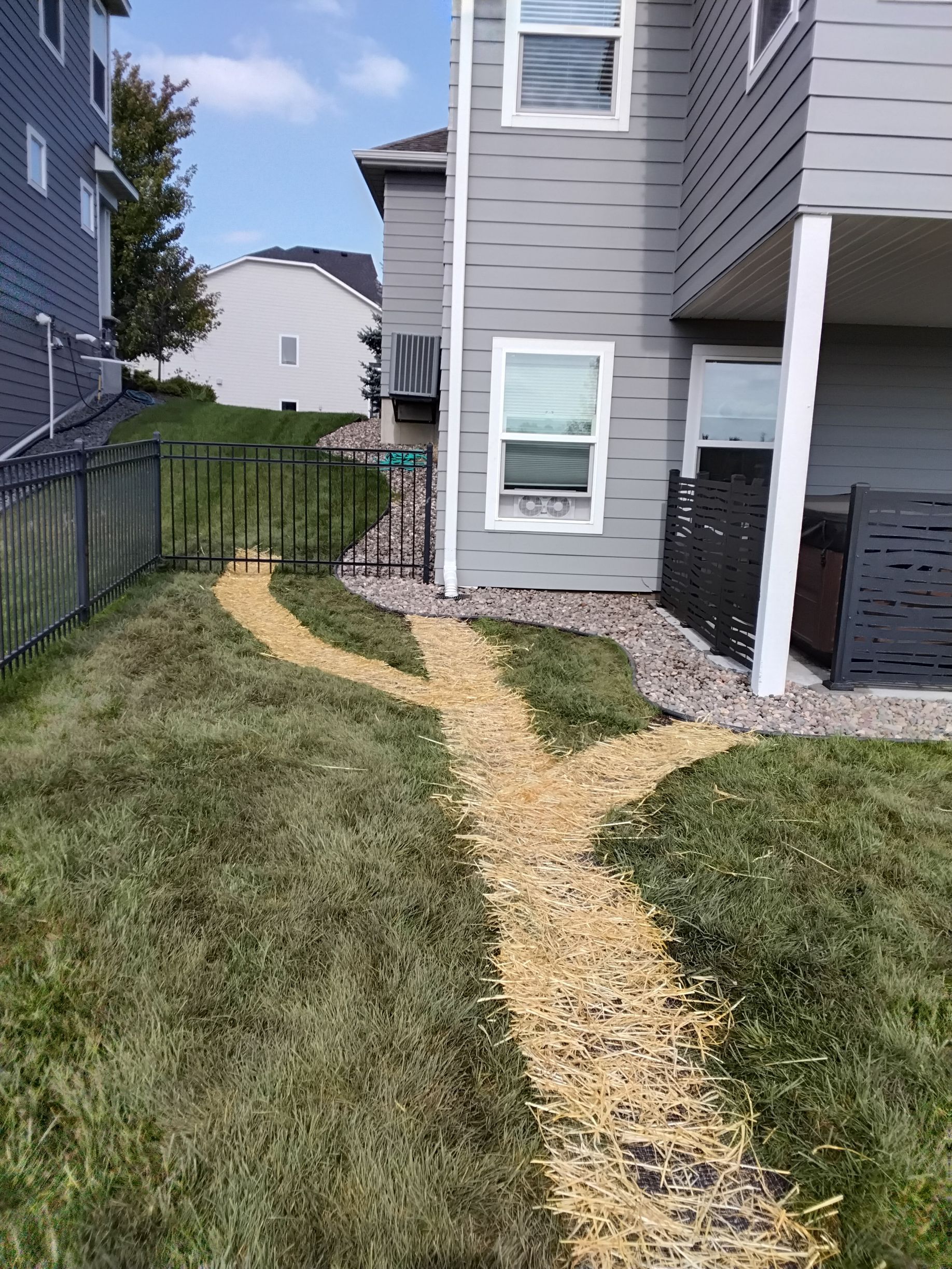 A mulched dirt path branches in two directions across a green lawn toward a metal gate and a house with grey siding.
