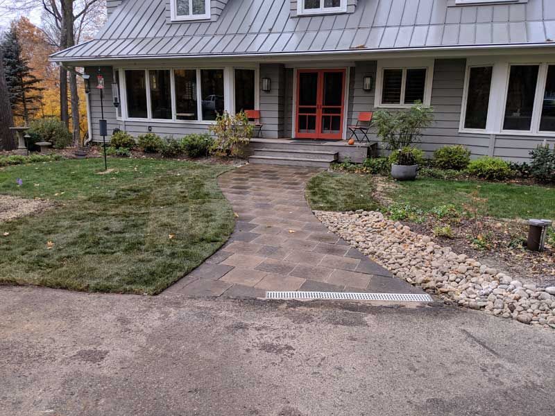 A gray house with a stone pathway leading to a red door, flanked by greenery and a rock border.
