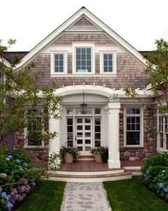 Stone and wood-shingled house with arched entry and white columns. Landscaped front yard with brick walkway.