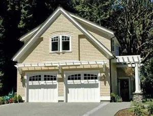 Tan two-car garage with white doors, pergola, and second-story windows under a gabled roof.