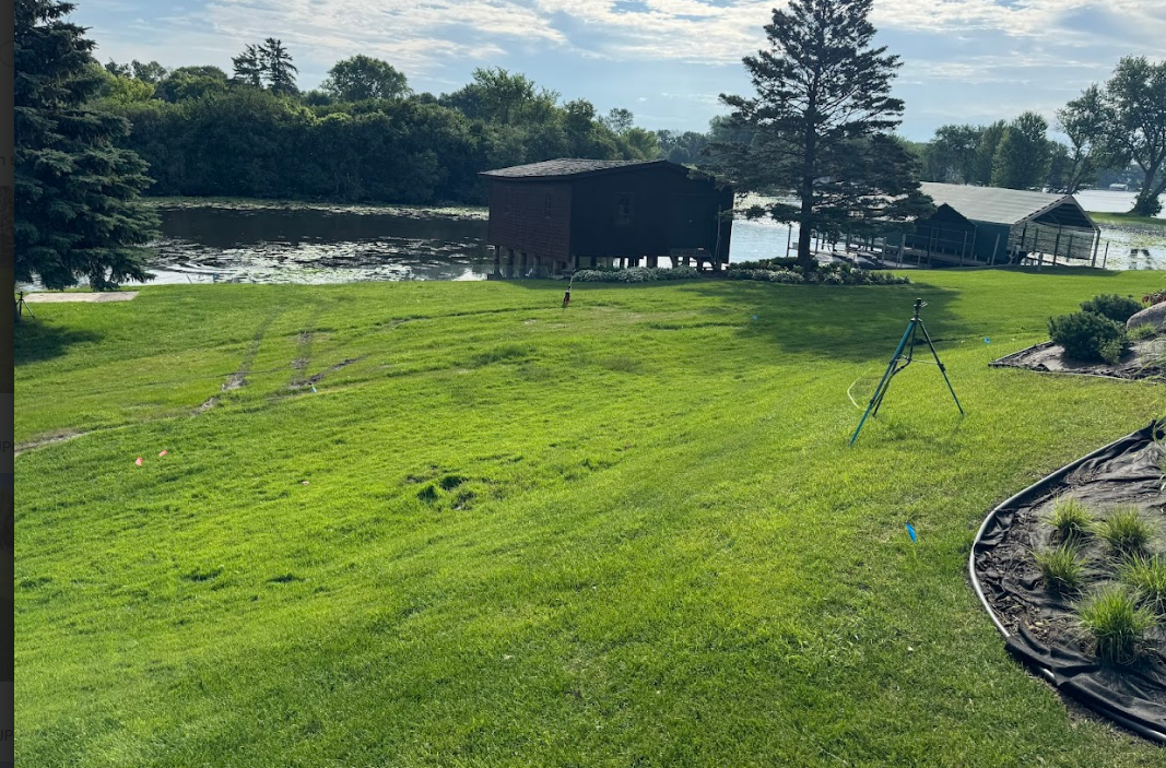A grassy backyard slopes toward a wooden boathouse and a river, with a tripod set up in the lawn.