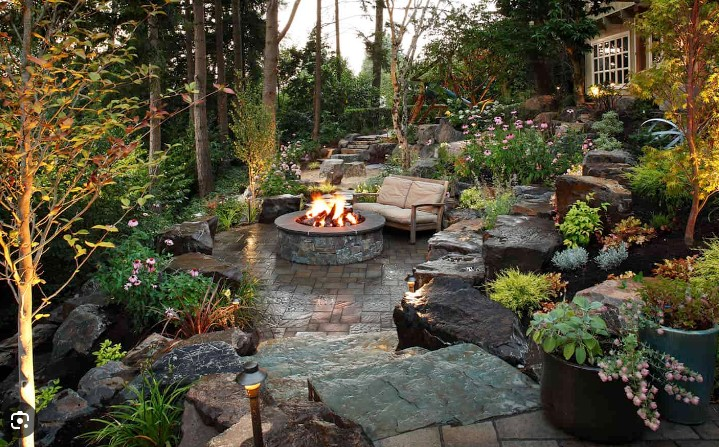 There is a fire pit in the middle of the garden with boulder wall and out cropping carved into backyard hill