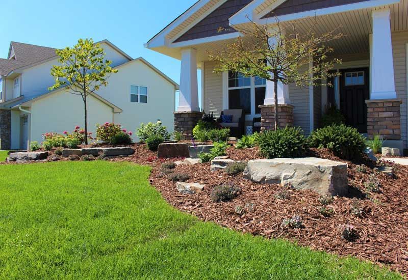 Well-manicured front yard with green grass, flower beds, and a beige house with white pillars.