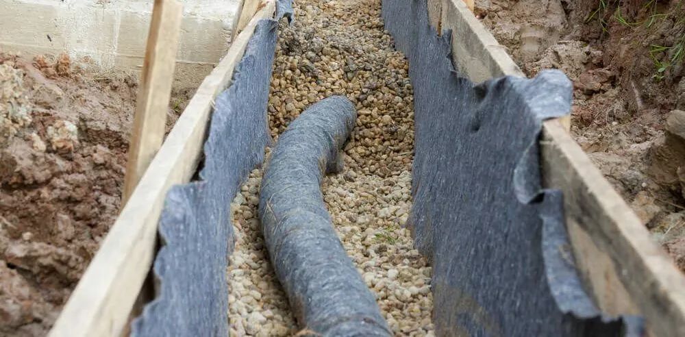 A French drain trench under construction with a fabric-wrapped pipe set in gravel between wooden forms.