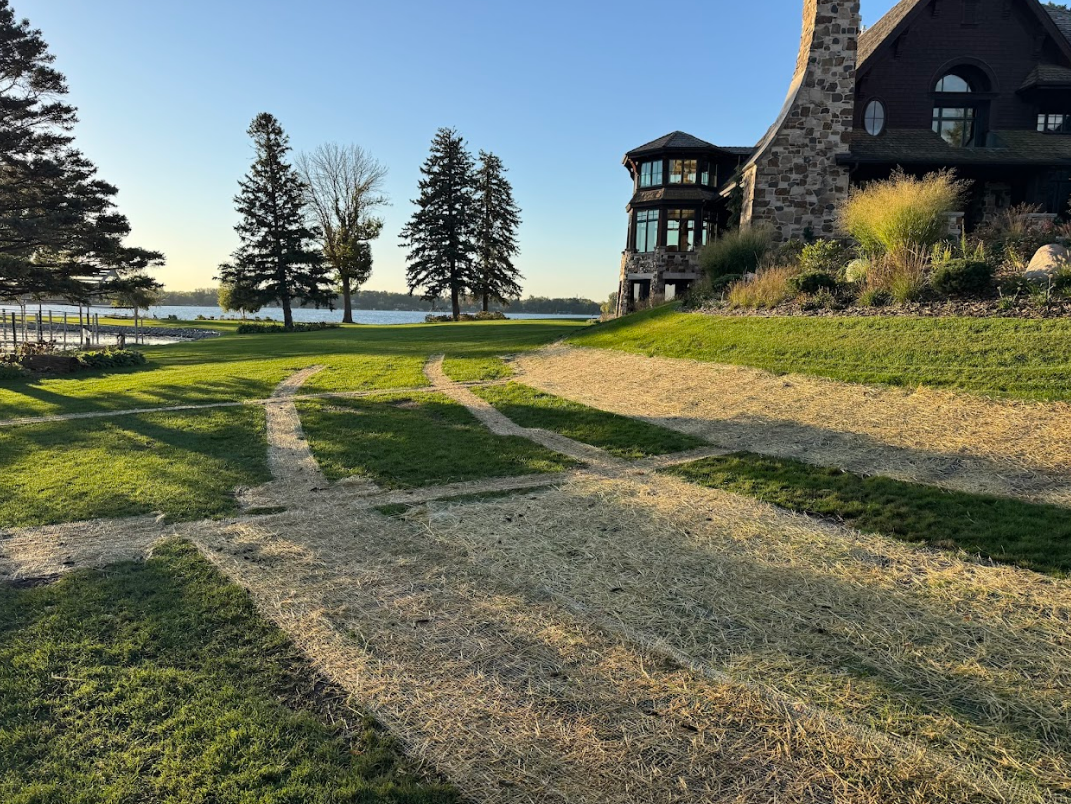 A stone house sits by a lake, with wide, dirt-filled drainage trenches cut into the grassy lawn in the foreground.