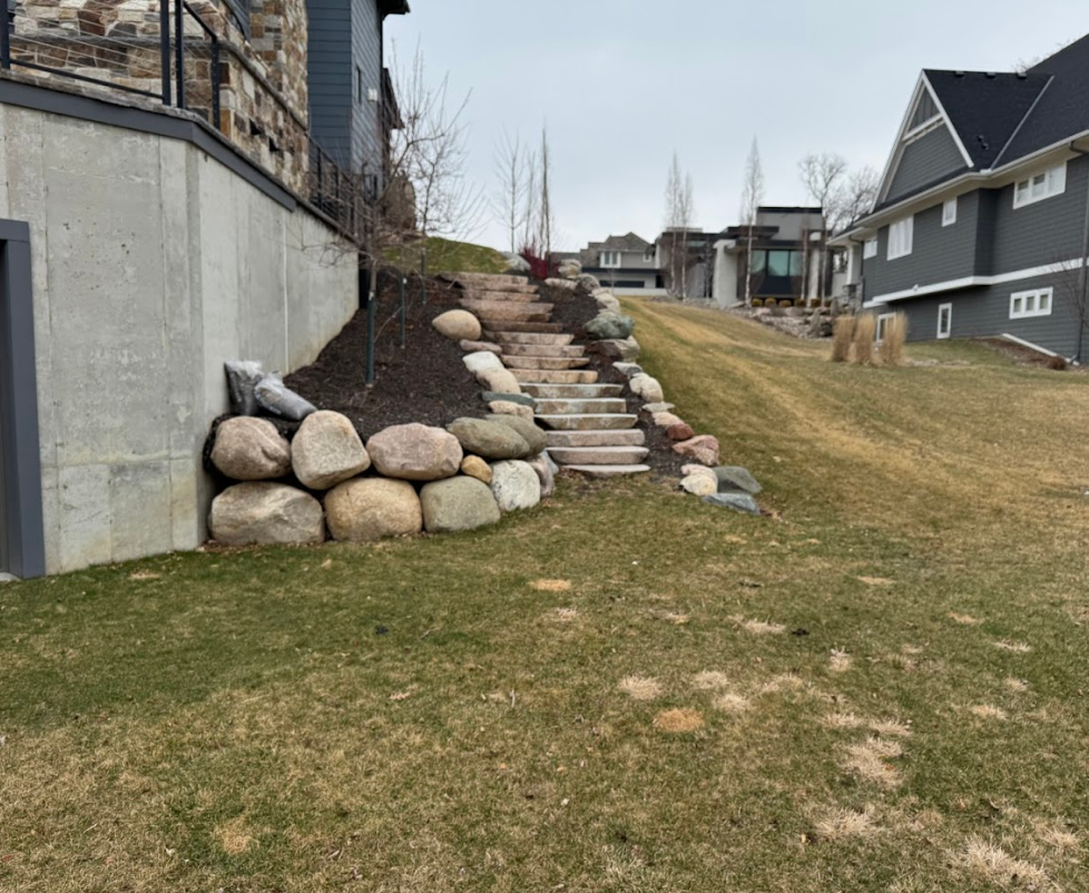 Stone steps lead up a grassy hill between two houses, with a large stone retaining wall on the left.