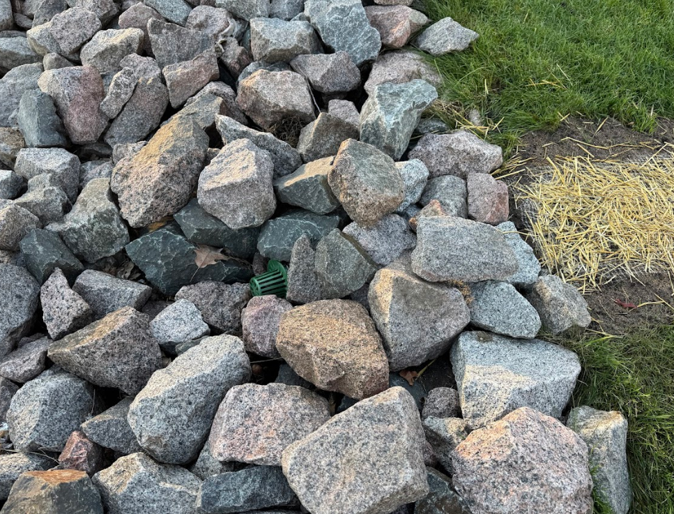 A close-up view of a pile of large, grey and tan granite rocks bordering a patch of grass.