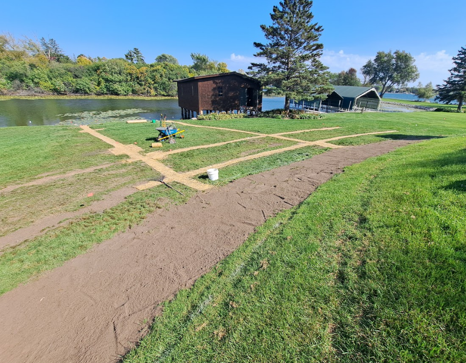 A grassy yard with freshly dug trenches and a dirt path leading toward a lakefront with two small wooden structures.