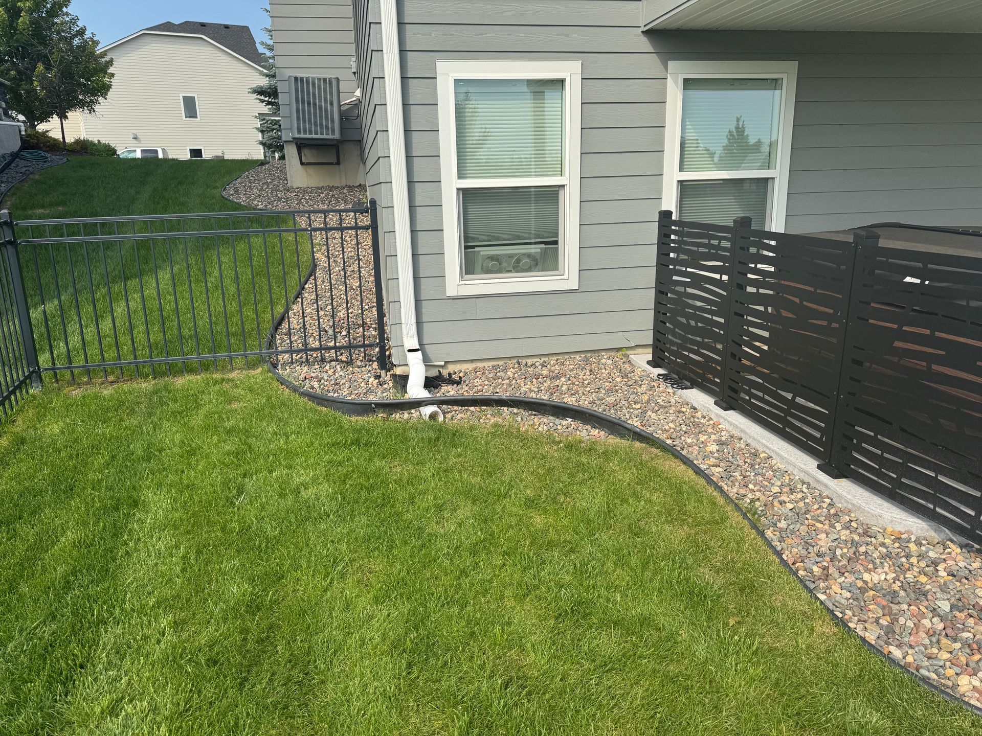 A residential exterior view featuring gray siding, two windows, a black privacy screen, and a gravel border along the lawn.