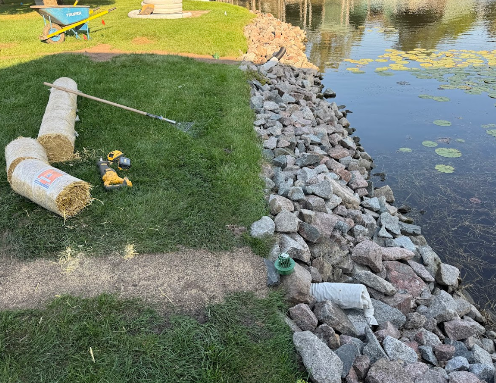 A lakeside embankment with rock riprap, straw rolls, and a drill on the grass, suggesting ongoing landscaping work.