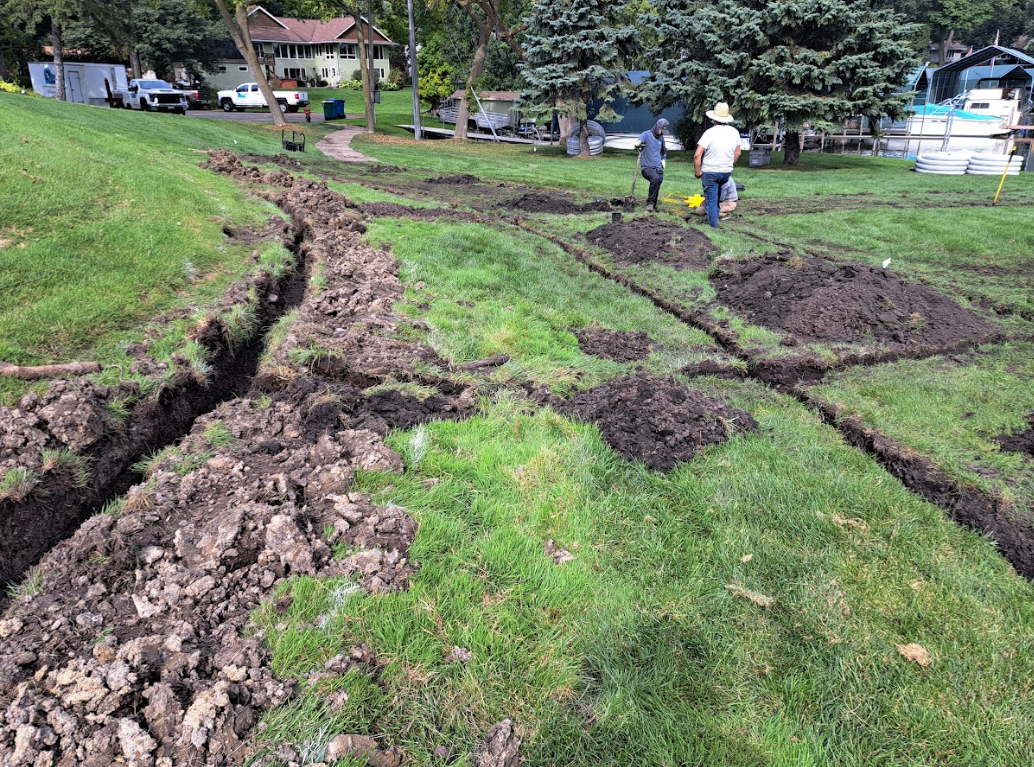 Two people work in a grassy yard, digging multiple narrow trenches into the soil for landscaping or utility installation.