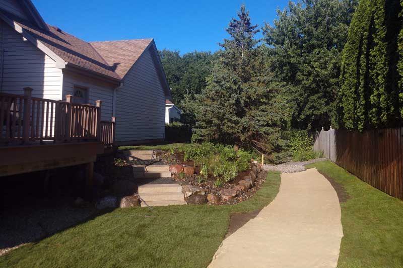 Backyard with a house, steps, path, and landscaping. Green grass, blue sky, and wooden fence.