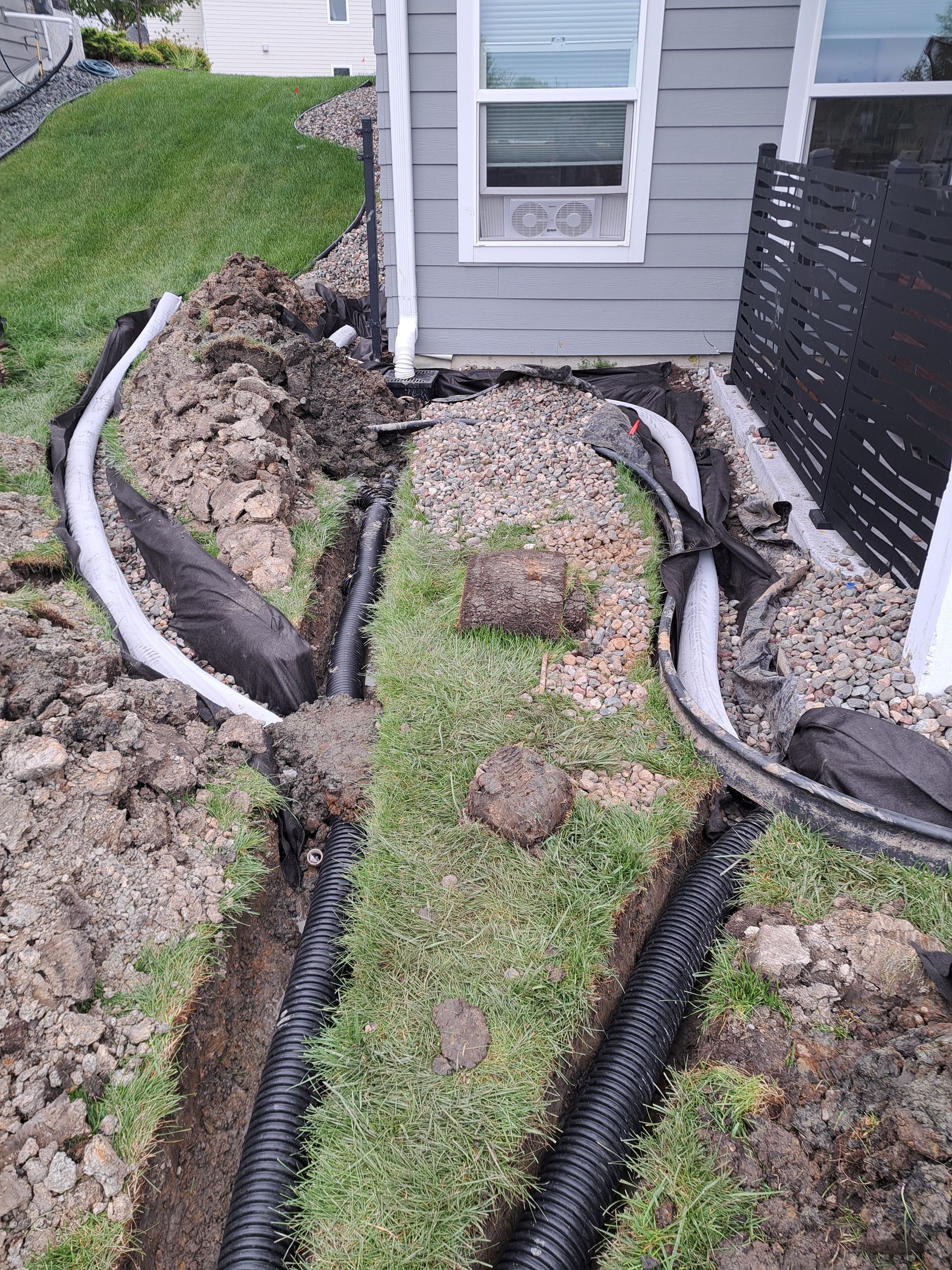 A side yard under construction featuring black corrugated drainage pipes installed in trenches next to the house foundation.
