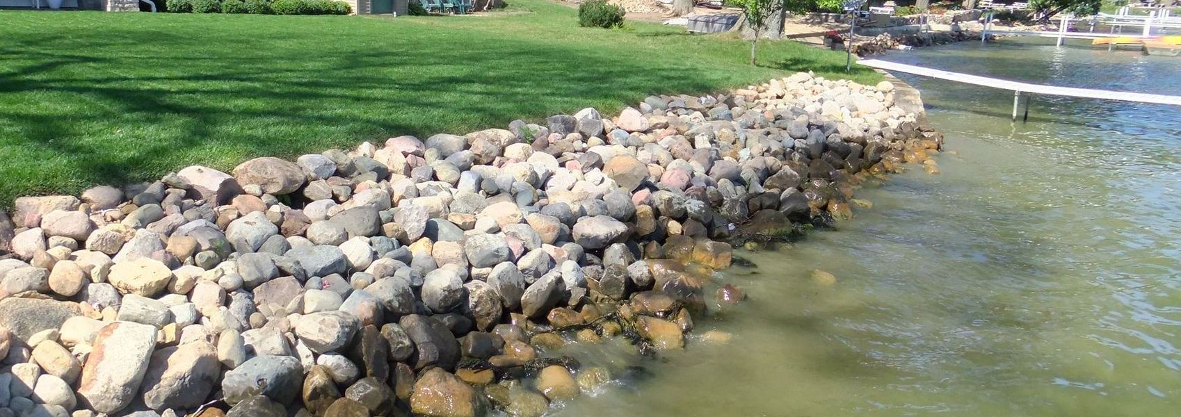 A rocky shoreline with a grassy bank leads to a body of water with a dock in the distance.