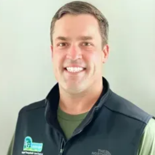 A smiling person wearing an olive-green shirt and a black branded vest against a plain light-colored background.