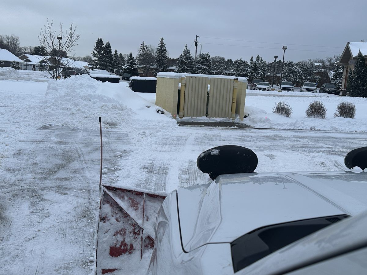 Plowed lot with snow banked beside a utility enclosure, seen from a plow truck