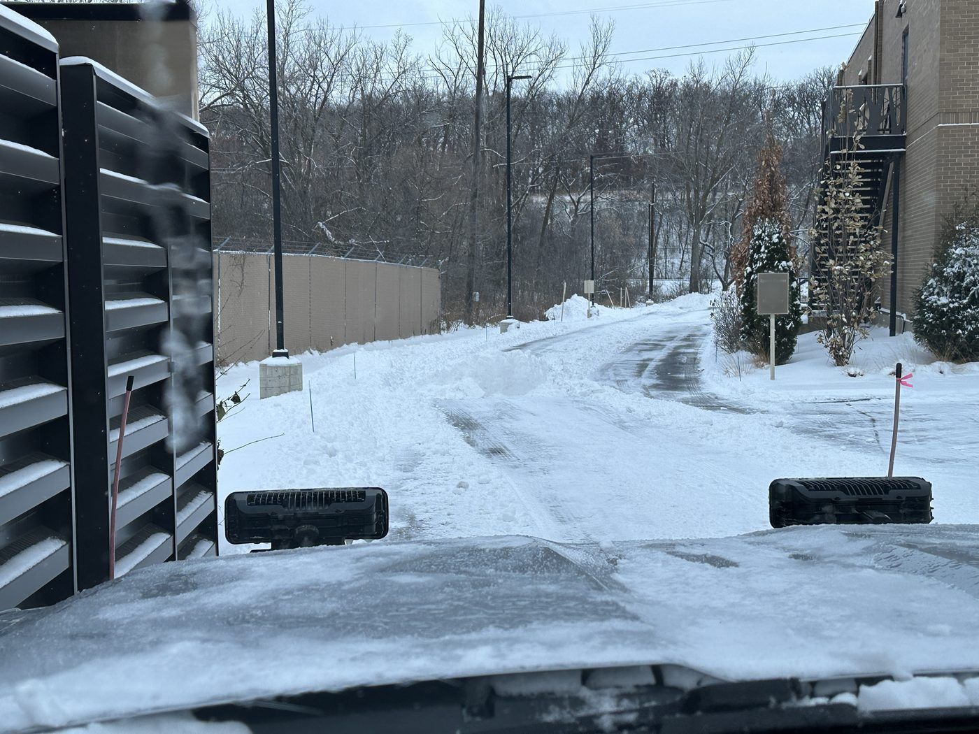 Plowed service drive beside a slatted enclosure and a brick building, seen from a plow truck