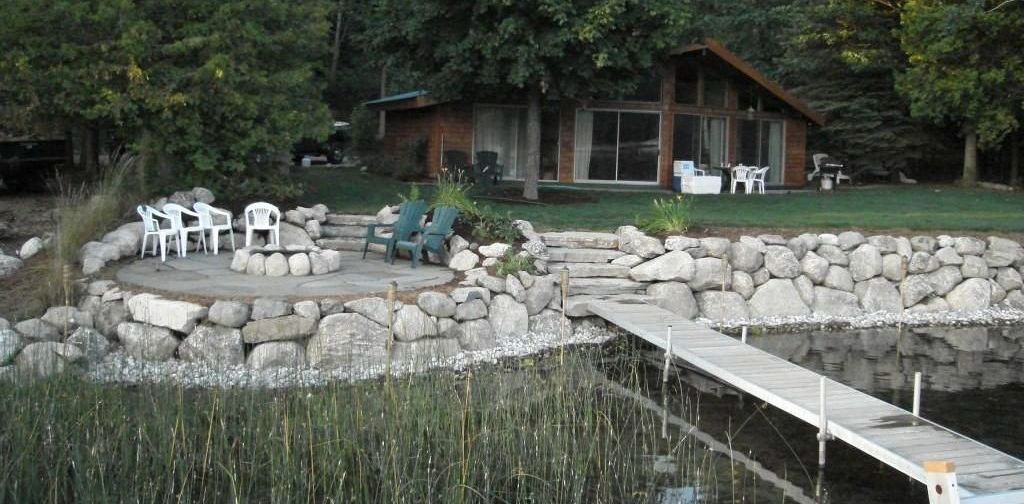 A cabin with a stone retaining wall, outdoor seating, and a dock extending into a lake with tall grass along the shore.