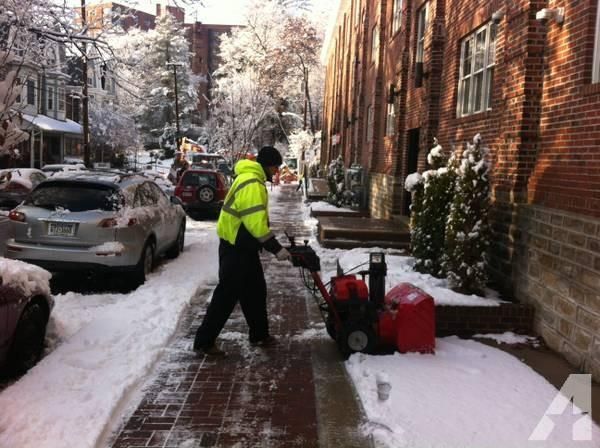 Person using a red snowblower on a snow-covered sidewalk, near a street with parked cars and brick buildings.