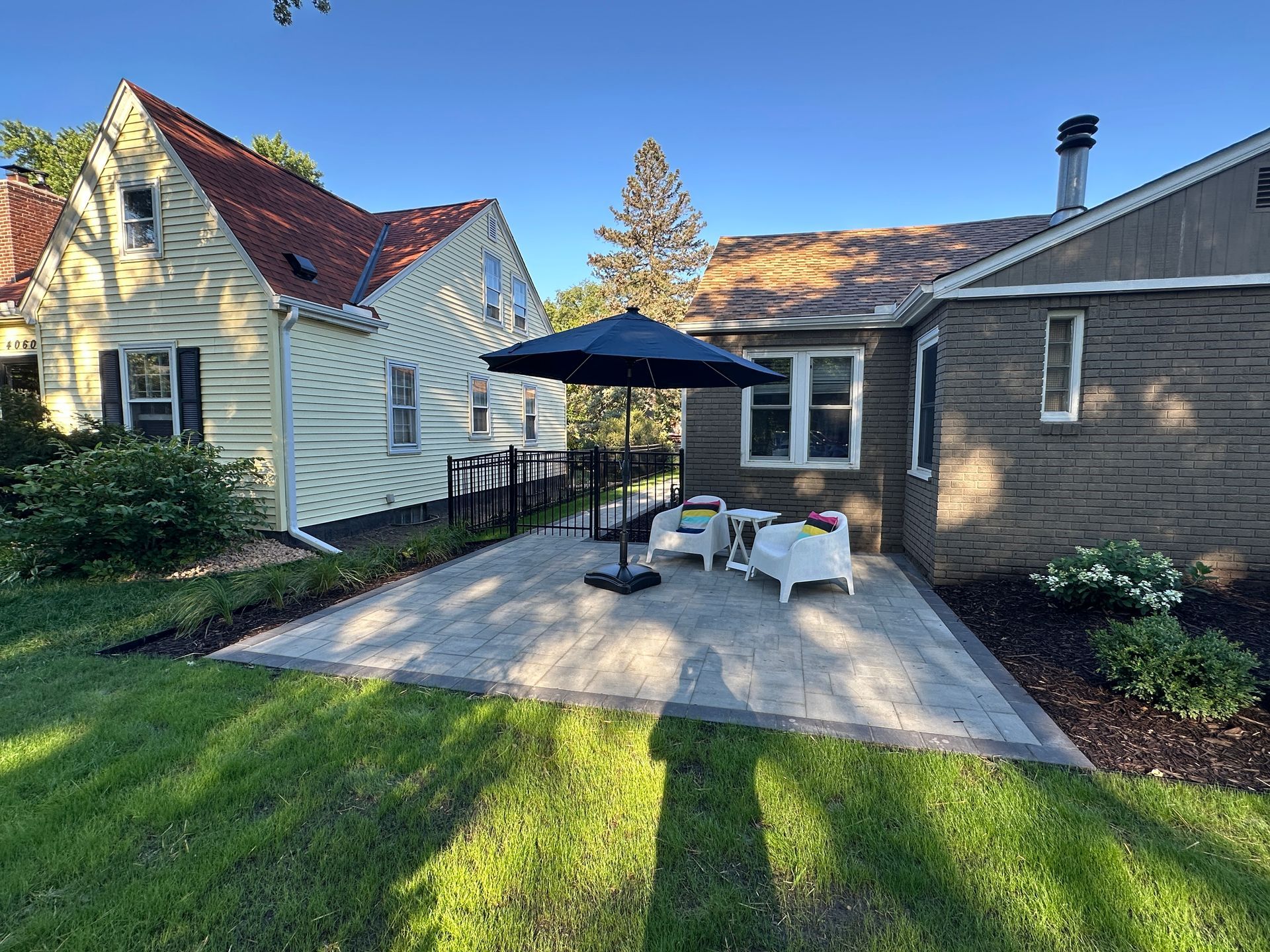 A stone patio with two white chairs and a blue umbrella situated between a yellow house and a tan brick house.