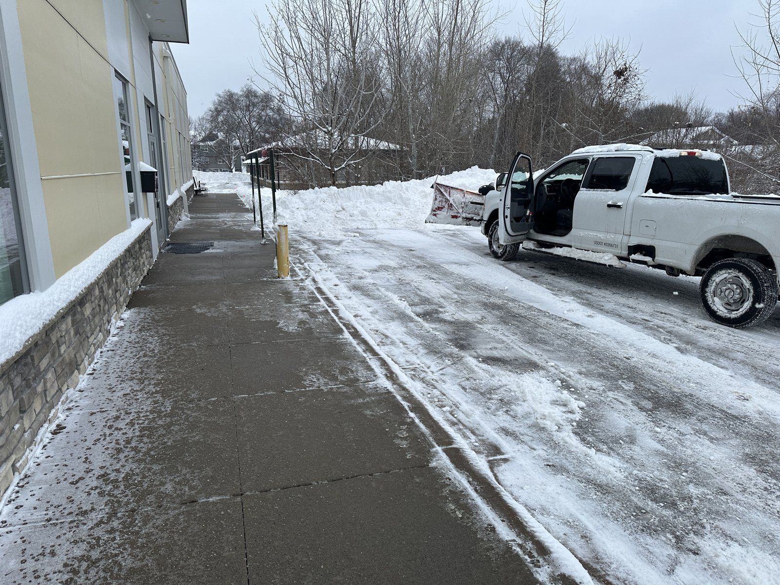 Walk along an office building with light snow left after plowing, and a plow truck in the drive