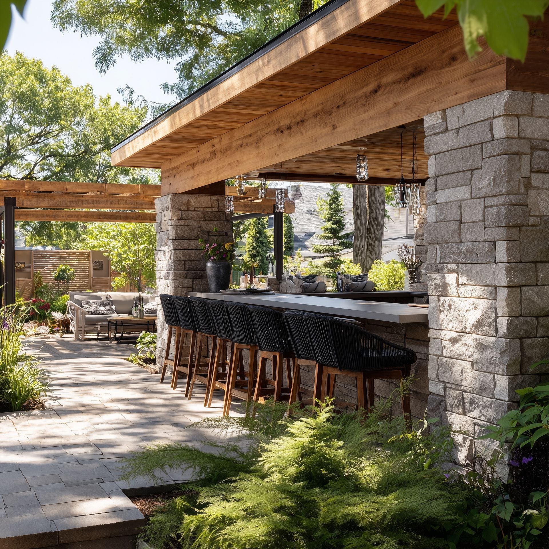 Outdoor bar with stone walls and wooden roof, lined with black woven bar stools, garden in foreground.