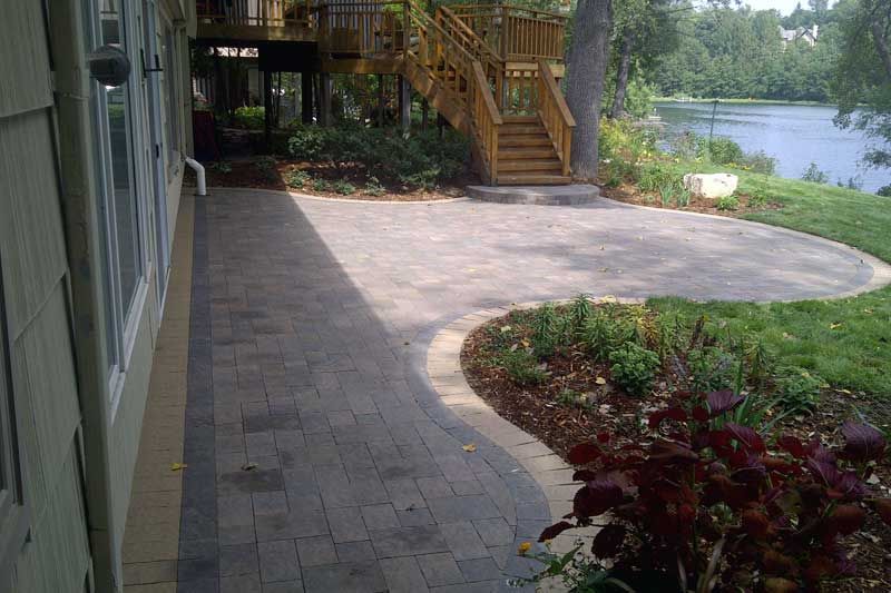Stone patio next to house, leading to a lawn and waterfront with wooden stairs.