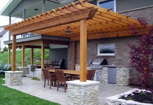 Patio with wooden pergola, dining set, built-in grill, and stone columns next to a brick building.