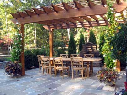 Wooden pergola over outdoor dining table and chairs on a stone patio, surrounded by plants and trees.
