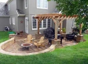 Backyard patio with wooden pergola, chairs, grill, and fire pit near a two-story house.