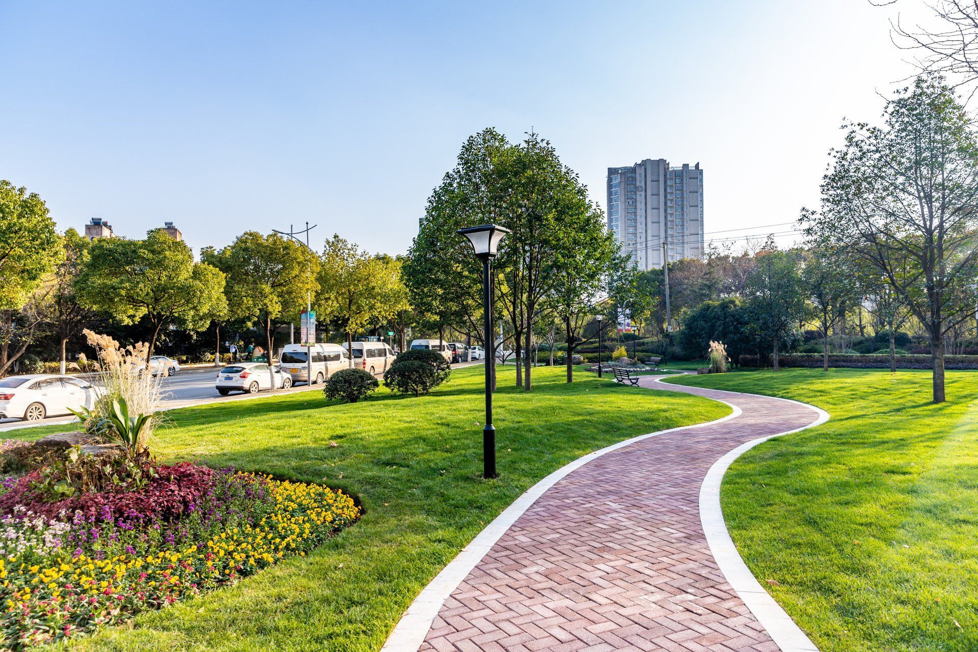 A brick path curves through a park with green grass, trees, and flowers. A city building is in the background.