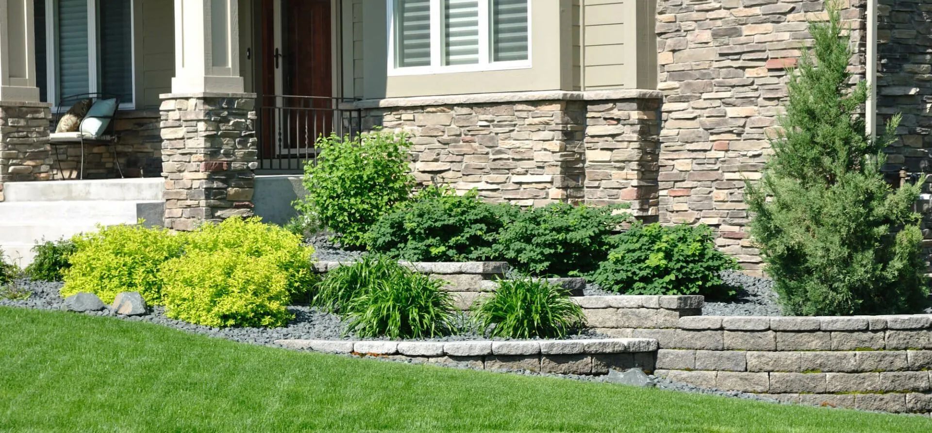 Front entrance of a house with stone siding, a tiered landscape wall, a small evergreen tree, and various shrubs.